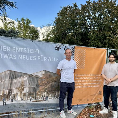 zwei Personen vor einem Bauzaunbanner das darüber Informiert was gebaut wird